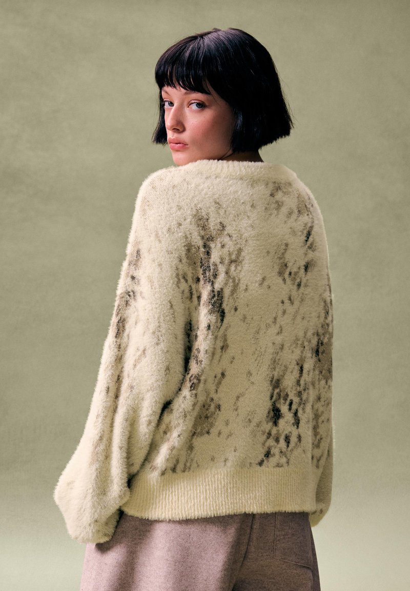 Młoda kobieta z krótkimi czarnymi włosami, ubrana w kremowy puszysty sweter z ciemnymi łatami, spoglądająca przez ramię na tle stonowanej zieleni.