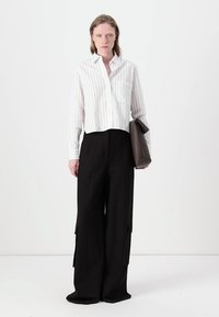 Camisetta cropped a righe bianche e grigie con maniche lunghe, abbinata a pantaloni larghi neri con tasche cargo. Borsa marrone in mano.