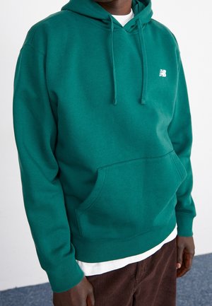 Homme portant un sweat à capuche bleu sarcelle avec une poche avant et un logo blanc, superposé sur une chemise blanche et un pantalon en velours côtelé marron.