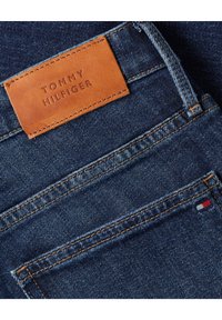 Poche arrière d’un jean en denim bleu foncé avec étiquette en cuir marron Tommy Hilfiger et petite languette de logo rouge, blanche et bleue.