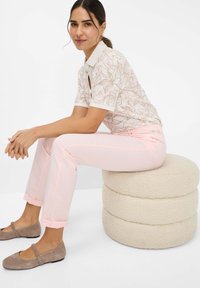 Een poloshirt met bloemenpatroon en een witte kraag, gecombineerd met lichtroze opgerolde broeken en taupe ballerina's, zittend op een ronde, gestructureerde kruk.