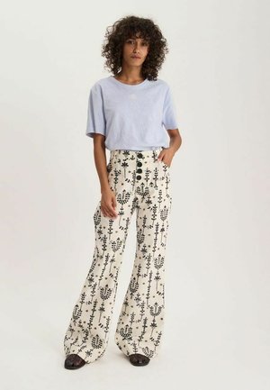 Femme aux cheveux bouclés portant un haut bleu clair à manches courtes et un pantalon large taille haute blanc avec imprimé floral noir et boutons.