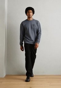 Sivi pamučni dukserica s okruglim izrezom i vezom, u kombinaciji s crnim ravnim hlačama i crnim cipelama na drvenom podu.