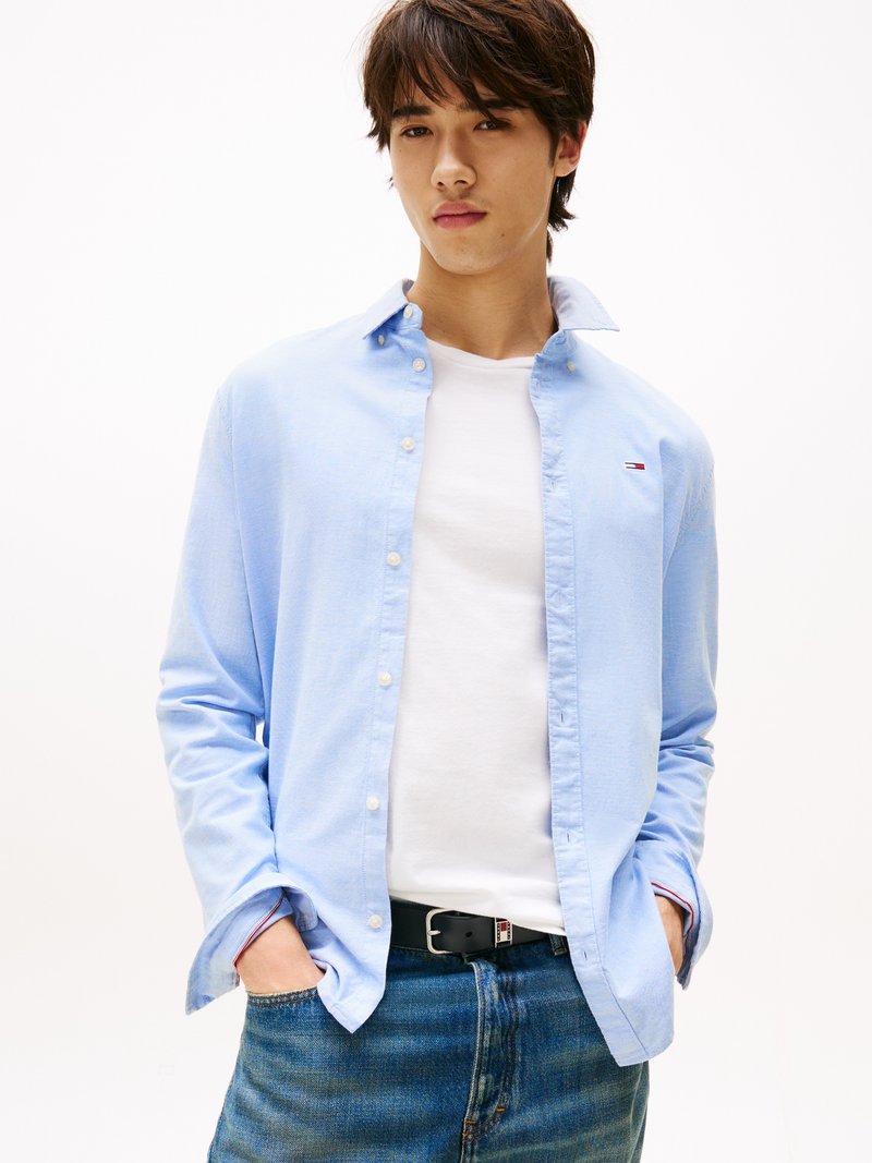 Camisa de botones azul claro con mangas largas, llevada sobre una camiseta blanca. Unos pantalones vaqueros oscuros y un cinturón negro completan el conjunto.