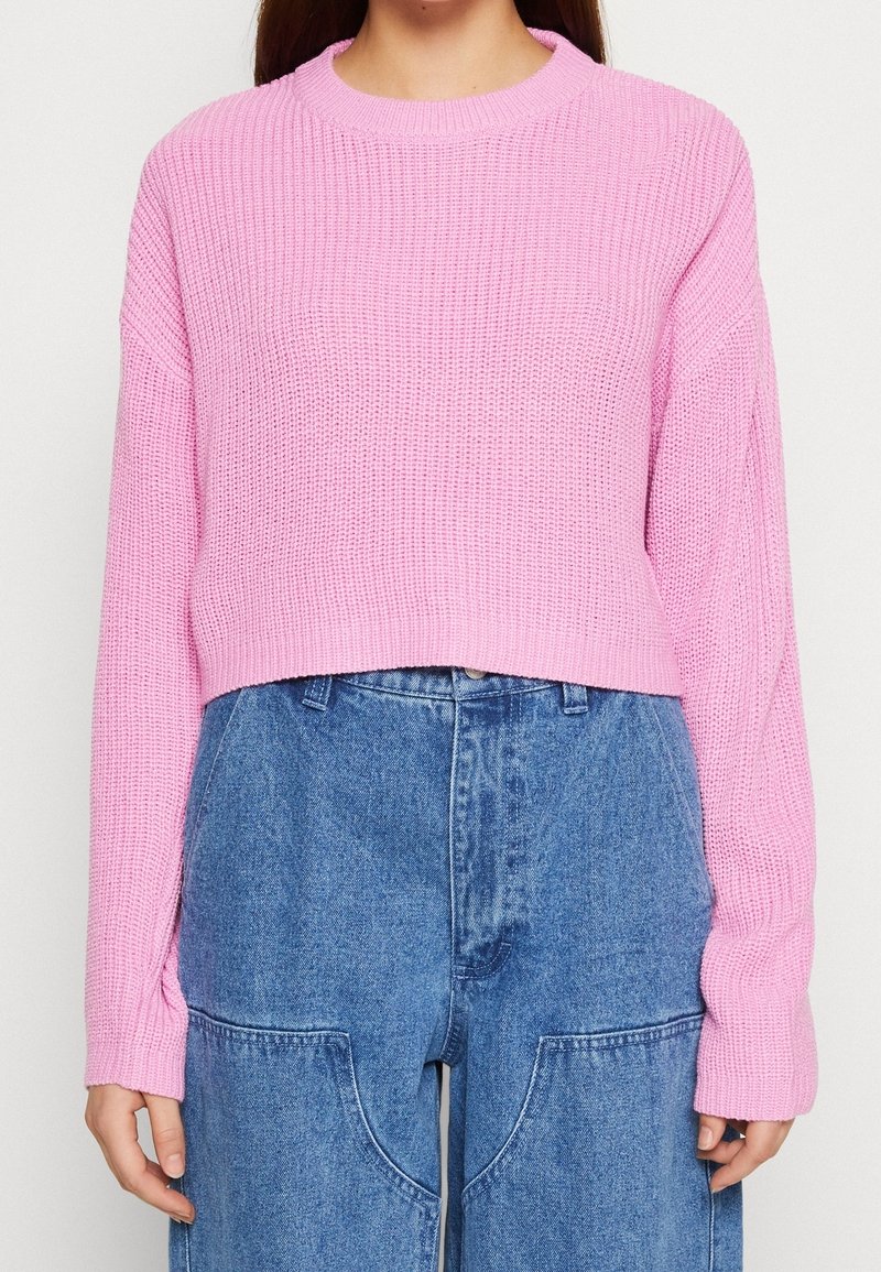 Rosa gestrickter, kürzerer Pullover mit strukturiertem Muster und langen Ärmeln, kombiniert mit hoch geschnittenen Jeanshosen mit großen Taschen.