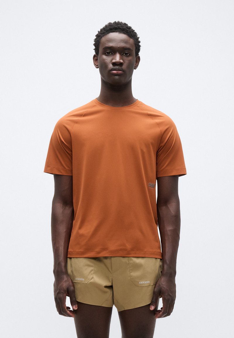 Πορτοκαλί αθλητικό μπλουζάκι με κοντά μανίκια και στρογγυλή λαιμόκοψη, με λείο ύφασμα και μικρό λογότυπο στην αριστερή πλευρά. Συνδυάζεται με μπεζ σορτς.