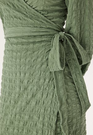 Olivgrön, texturerad omlottklänning med skrynkligt tyg och sidoknytning. Har ett subtilt rutmönster och långa ärmar.