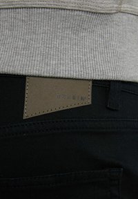 Jeans negros con una cinturilla acanalada gris texturizada. Una etiqueta de cuero marrón con "CHASIN'" en relieve en negro está adjunta en la parte posterior.