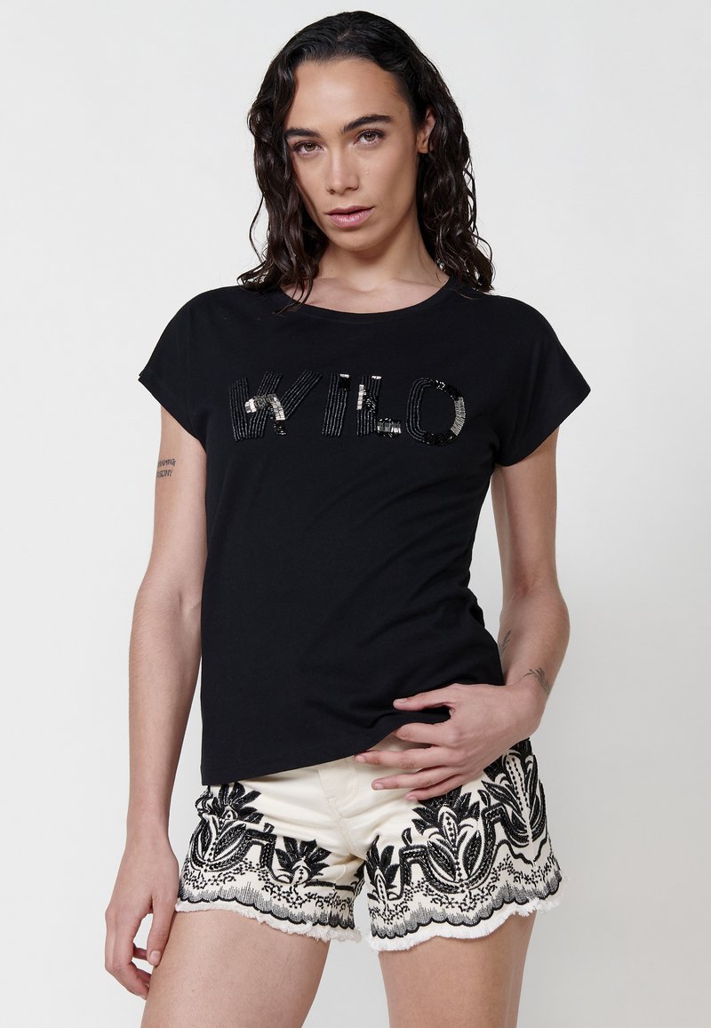 Camiseta negra de algodón con mangas cortas enrolladas, que presenta un texto de lentejuelas 'WILO' en la parte frontal. Combinada con pantalones cortos crema con bordado floral negro.