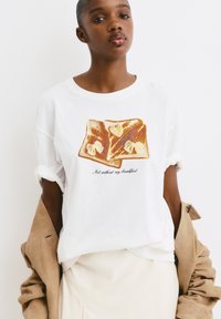 Witte katoenen t-shirt met een illustratie van twee boterhammen met boter en een geschreven tekst die zegt "Niet zonder mijn ontbijt."