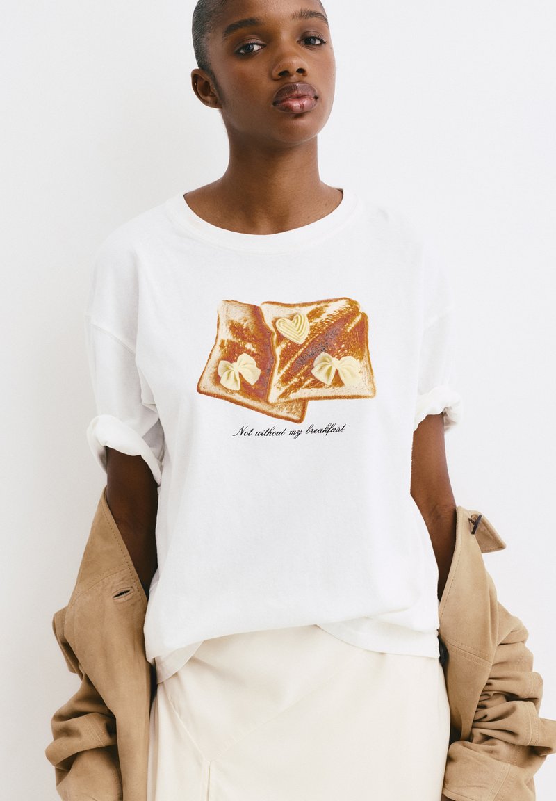 Witte katoenen t-shirt met een illustratie van twee boterhammen met boter en een geschreven tekst die zegt "Niet zonder mijn ontbijt."