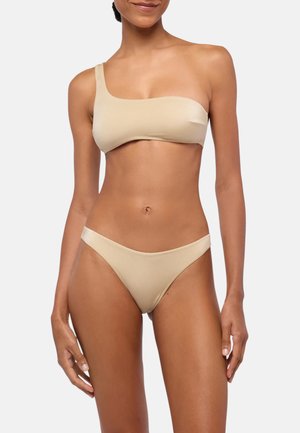 Donna che indossa un top bikini beige monospalla e slip coordinati su uno sfondo chiaro semplice.