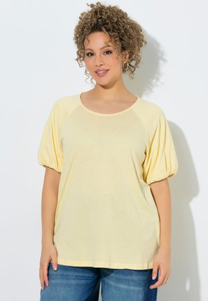 Femme aux cheveux bouclés portant un haut jaune à manches bouffantes et un jean bleu, debout devant un fond clair uni.