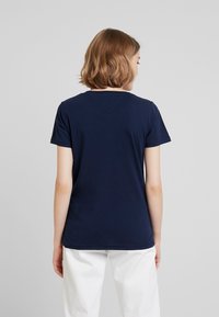 Námořnická modrá tričková halenka s krátkým rukávem, kulatým výstřihem, hladkou texturou a detailem ve tvaru V na zadní straně. Nosená s bílými kalhotami.