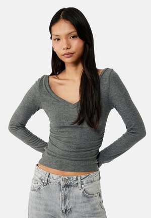 Aziatische vrouw met lang zwart haar, gekleed in een aansluitende grijze geribde V-hals trui en lichtgrijze spijkerbroek, staand met handen in de zij.