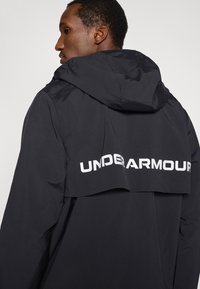 Μαύρο αδιάβροχο μπουφάν με κουκούλα· διαθέτει λευκό λογότυπο "UNDERARMOUR" στην πλάτη· ομαλή υφή και αθλητικός σχεδιασμός.