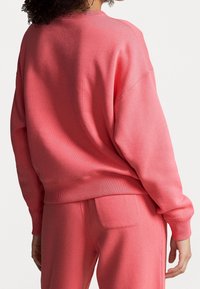 Rosa sweatshirt med avslappnad passform, ribbade ärmslut och fåll, tillsammans med matchande rosa byxor som har en enda bakficka. Slät textur.