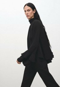 Top negro de manga larga con cuello alto, que presenta tela drapeada y puños acampanados. Combinado con pantalones negros a juego, de diseño elegante y minimalista.