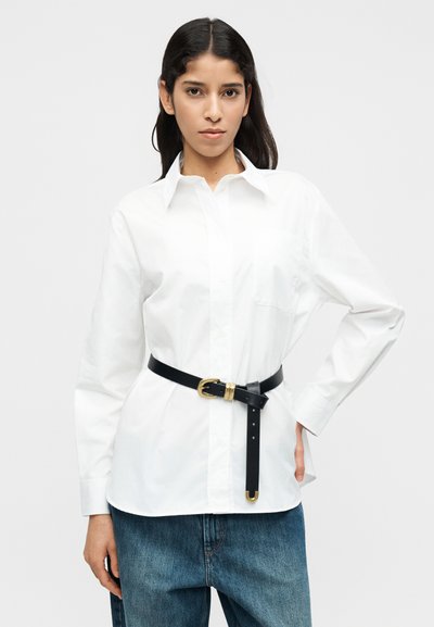 Λευκό πουκάμισο με κουμπιά, με μυτερό γιακά και μία τσέπη στο στήθος, εφαρμοστό στη μέση με μαύρη ζώνη που έχει χρυσή αγκράφα.