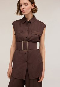 Camicia marrone con cintura, finitura testurizzata, maniche corte, chiusura a bottoni e due tasche sul petto, con una larga fibbia dorata rettangolare.