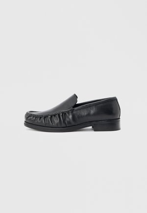 Black leather men's slip-on loafer with rounded moc toe, ruched detailing, and low block heel on white background.