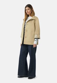 Beżowa kurtka w stylu cropped z wysokim kołnierzem, metalowymi guzikami i strukturalnymi mankietami, w połączeniu z ciemnymi spodniami dzwonami i swetrem w paski.