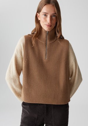 Eine Frau trägt einen beige gerippten ärmellosen Strickpullover mit einem hochstehenden Reißverschlusskragen über einem cremefarbenen Langarmpullover und dunklen Hosen.