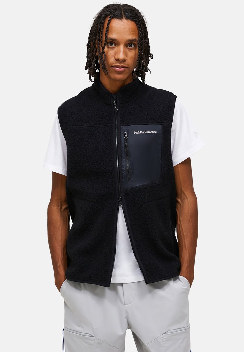 Νεαρός άνδρας με κοτσίδες φοράει μαύρο φλις γιλέκο με φερμουάρ πάνω από λευκό μπλουζάκι και ανοιχτόχρωμο γκρι παντελόνι, στέκεται με τα χέρια στις τσέπες.