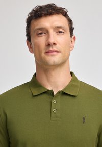 Grön polo med mjukt tyg som har en treknapps-knäppning och en liten broderad logotyp på vänster bröst.