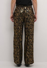 Nainen, joka pukeutunut mustiin leveälahkeisiin housuihin, joissa on kultainen metallinen abstrakti kuvio, seisoo käsivarret rentoina sivuilla yksiväristä taustaa vasten.