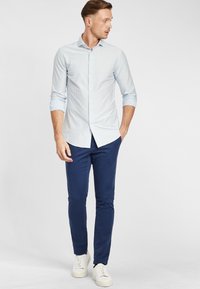 Camicia a maniche lunghe a righe blu chiaro con colletto button-down, abbinata a pantaloni slim-fit blu navy e sneakers bianche. Design minimalista.