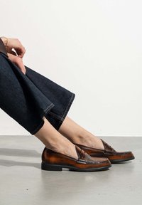 Brown leather loafers with a textured finish, featuring decorative stitching and a low heel. Paired with ankle-length dark denim pants.