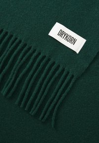 Écharpe en laine vert foncé avec des bords à franges et une étiquette blanche portant le texte "DRYKORN". La texture semble douce et chaude.