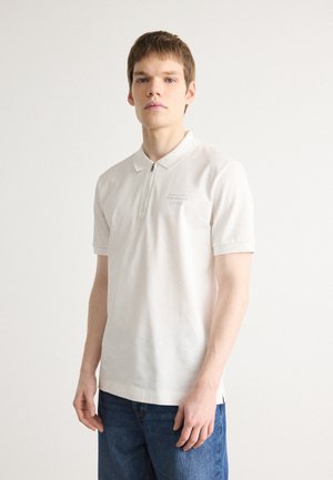 Polo bianco con zip corta e colletto, realizzato in tessuto morbido. Presenta un sottile logo "BOSS" grigio sul petto, abbinato a jeans blu.