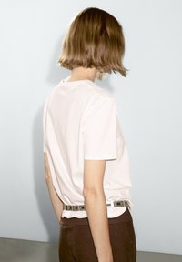 T-shirt di cotone rosa chiaro a maniche corte indossato con pantaloni marroni e una cintura con fibbia argentata con dettagli quadrati.
