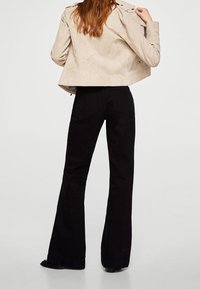 Donna con capelli lunghi indossa una giacca corta beige e pantaloni neri a vita alta e svasati, in piedi di spalle con una mano sul colletto.