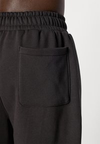 Nærbillede af sorte sweatpants med elastisk talje og en firkantet patchlomme på højre hofte mod en ensfarvet baggrund.
