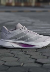 Hellgrauer Adidas Laufschuh mit drei silbernen Streifen, gepolsterte Sohle mit lila Detail, auf sechseckigem Steinpflaster.