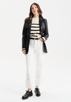 Blazer di pelle nera su un pullover a righe bianche e nere, abbinato a pantaloni bianchi a gamba larga e scarpe nere con lacci.