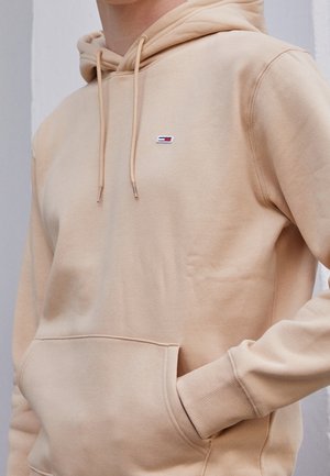 Personne portant un sweat à capuche beige avec un petit logo rectangulaire rouge, blanc et bleu sur la poitrine et la main partiellement dans la poche avant.