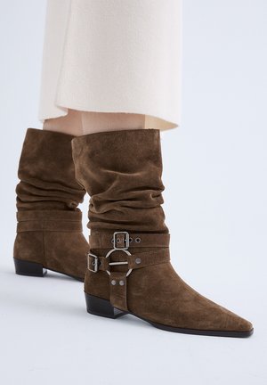 Botas marrones de ante hasta media pantorrilla con diseño holgado, tiras decorativas, anillas de metal y tacones bajos tipo bloque, usadas con una falda de color crema.