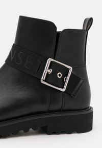 Černá kožená kotníková bota s robustní podrážkou a texturovaným černým páskem s kovovou sponou stříbrné barvy a vyrytým nápisem.