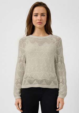 Femme portant un pull à manches longues beige clair avec des motifs en maille ajourée complexes, associé à un pantalon noir, debout devant un fond uni.