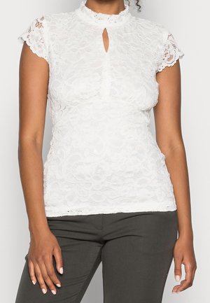 Mujer con una blusa blanca de encaje de manga corta con cuello simulado en forma de ondas y detalle con abertura, combinada con pantalones oscuros.