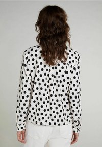 Blusa a pois bianca e nera con colletto, polsini con bottone e schiena plissettata. Realizzata in un tessuto liscio con una leggera lucentezza.