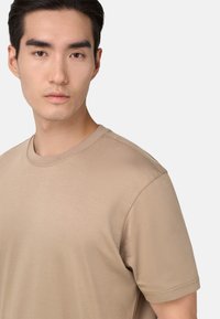 Béžové tričko s krátkým rukávem vyrobené z měkké látky. Má kulatý výstřih a volný střih. Viditelné švy na rukávech.