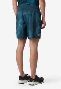 Men's athletic shorts in teal with a water-like pattern. Lightweight fabric and elastic waistband. Paired with black running shoes.