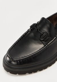 Mocassins en cuir noir avec un bout arrondi, des garnitures décoratives et une semelle en caoutchouc épaisse avec un motif texturé.