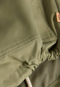 Tuvaļskats uz olīvzaļas auduma ar izteiktu šuvumu un elastīgu krokojumu, parādot tekstūru un nelielu bēša auduma etiķeti ar melnu tekstu.