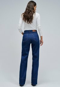Mujer con cabello largo y castaño de espaldas, vestida con una blusa blanca de manga larga y unos vaqueros oscuros de tiro alto con dobladillos.
